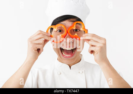 Emozionato chef asiatico che indossano uniformi isolato permanente su sfondo bianco, divertendosi, azienda di capsicum a fette Foto Stock