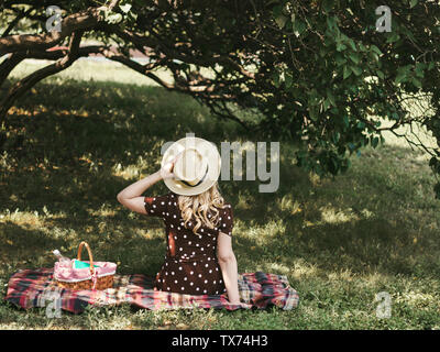 Picnic sulla vacanza estiva foto con spazio copia di una giovane ragazza bionda in un cappello di paglia è a riposo nel giardino in una giornata soleggiata vista posteriore Foto Stock