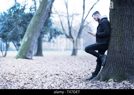 Uno stile retrò foto di un giovane hipster uomo appoggiato a un albero e la lettura di un libro nel parco. Foto Stock