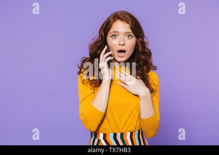 Ritratto di un piuttosto confuso redheaded giovane donna in piedi isolato su sfondo viola, utilizzando il telefono cellulare Foto Stock