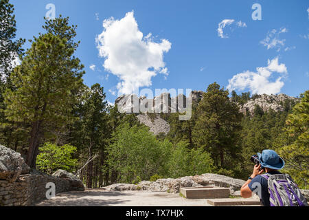 Un giovane che sta alla base della montagna per scattare una foto del monte Rushmore con alberi di pino e cielo blu. Foto Stock