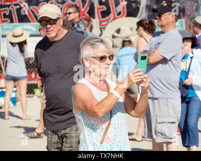Un senior donna in occhiali da sole scatta una foto con un telefono cellulare al 2019 Texas Sandfest in Port Aransas, Texas, Stati Uniti d'America. Foto Stock