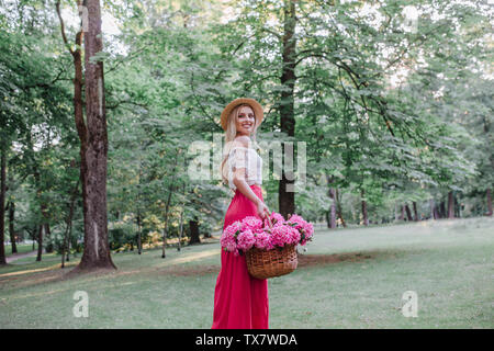 Bella ragazza in Vintage dress e cappello di paglia holding peonie rosa nel cesto rustico in estate park Foto Stock