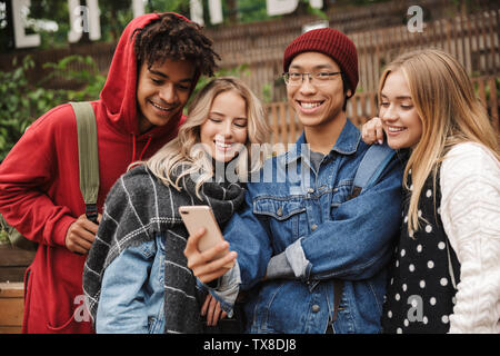 Se il gruppo di allegro multietnica amici adolescenti spendere tempo di divertimento insieme all'aperto, in piedi, utilizzando i telefoni cellulari Foto Stock