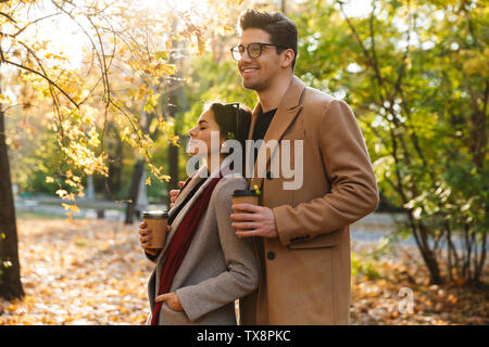 Ritratto di coppia romantica a bere il caffè da asporto da bicchieri di carta e abbracciando mentre passeggiate nel parco di autunno Foto Stock