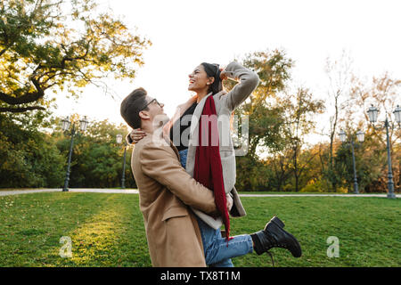 Immagine di elegante giovane uomo che porta la donna a mani e sorridere mentre passeggiate nel parco di autunno Foto Stock