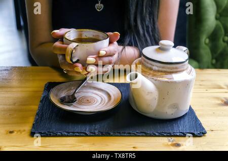 Persona che tiene una tazza bianca di tè di ceramica piena di tè accanto ad una pentola da tè allestita su un legno tabella Foto Stock