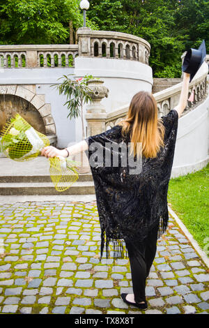 Felice studente ragazza con black robe accademico tiene nelle sue mani il tappo accademico e il mazzetto di fiori nel giorno d'inizio Foto Stock