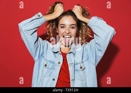 Ritratto di un allegro elegante giovane donna indossando giacca denim in piedi isolato su sfondo rosso, posa, con la lingua fuori Foto Stock