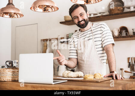 Ritratto di giovane uomo bruna 30s indossando il grembiule utilizzando computer portatile durante la cottura e rendendo la pasta fatta in casa in cucina a casa Foto Stock