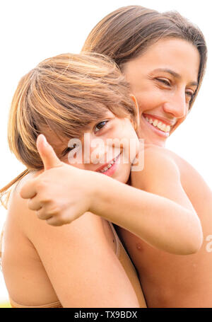 Ritratto di ragazzo dando un pollice in alto e guardando nella telecamera mentre viene amorevolmente abbracciato da sua madre Foto Stock