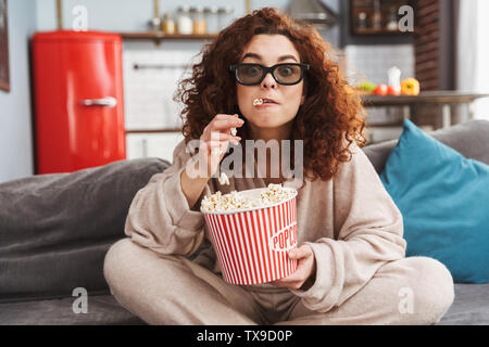 Foto di soggetti di razza caucasica giovane donna che indossa gli occhiali 3D mangiare popcorn mentre è seduto sul divano in appartamento Foto Stock