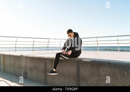 Foto di elegante guy 20s in nero legatura tuta sua lacci delle scarpe mentre è in esecuzione dal mare lungo il molo durante la mattina allenamento Foto Stock