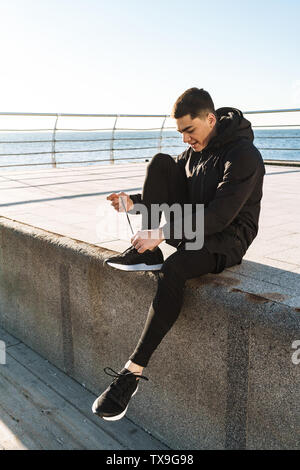 Foto di felice ragazzo 20s in nero legatura tuta sua lacci delle scarpe mentre è in esecuzione dal mare lungo il molo durante la mattina allenamento Foto Stock