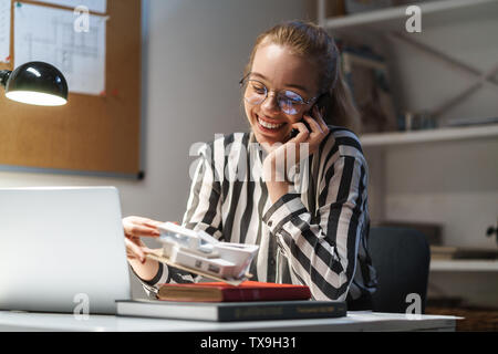 Foto di ottimista donna caucasica architetto indossando occhiali azienda cellulare e modello di casa mentre è seduto al posto di lavoro Foto Stock