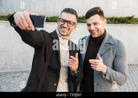 Due uomini belli che indossano tuniche di trascorrere del tempo all'aperto, a bere il caffè da asporto, camminare, parlare, prendendo un selfie Foto Stock