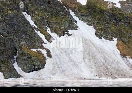 Vicino la vista attraverso gli occhi del lago, uno dei sette laghi di Rila, con colorati, ghiaccio rotto in corrispondenza di una scogliera rocciosa coperta di neve Foto Stock