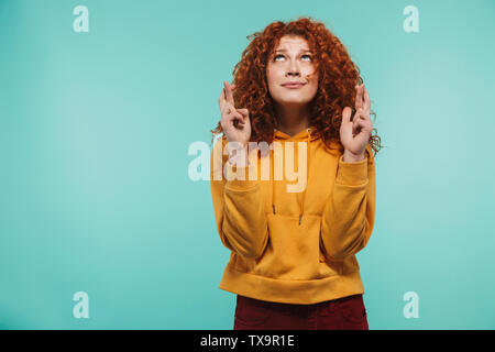 Immagine di donna affascinante 20s con ricci capelli allo zenzero mantenendo le dita incrociate e mendicando per buona fortuna isolato su sfondo blu Foto Stock