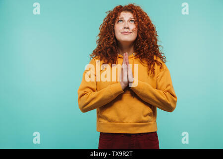 Immagine di donna gorgeous 20s con ricci capelli allo zenzero mantenendo le palme insieme e di pregare per la buona fortuna isolato su sfondo blu Foto Stock