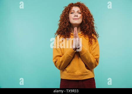 Immagine di donna attraente 20s con ricci capelli allo zenzero mantenendo le palme insieme e di pregare per la buona fortuna isolato su sfondo blu Foto Stock