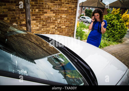 Giovane donna sul telefono cellulare arrabbiato con parabrezza rotto sulla sua automobile. Parabrezza è un chip e di una fessura in un angolo. Foto Stock