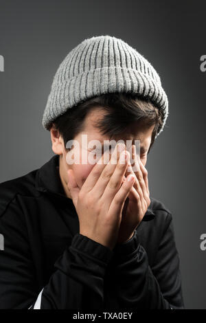 Ragazzo di 11 anni in tuta nera e grigia wooly hat piangendo con il suo volto nelle sue mani Foto Stock