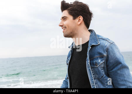 Attraente giovane uomo indossando giacca denim passeggiate in spiaggia Foto Stock