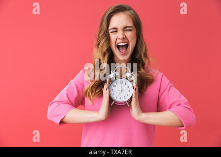 Bella scioccato confuso giovane ragazza di indossare abiti casual in piedi isolato su sfondo rosa, che mostra orologio sveglia Foto Stock