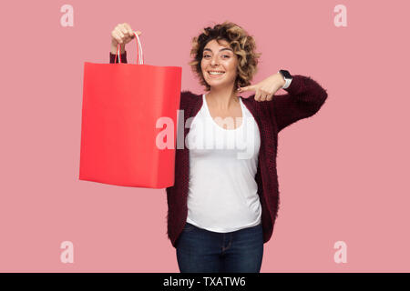 Allegro bella giovane donna in maglietta blu con curlty acconciatura permanente e puntare il dito di borse per lo shopping e il sorriso toothy, guardando la fotocamera. Foto Stock