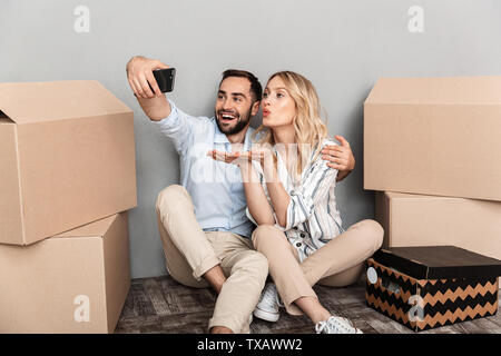 Foto di coppia attraente posto a sedere vicino a scatole di cartone e tenendo selfie foto sul cellulare isolate su muro grigio Foto Stock