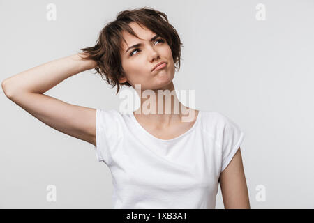 Ritratto di giovane donna con capelli bruni corti in base t-shirt accigliata e guardando verso l'alto isolato su sfondo bianco Foto Stock