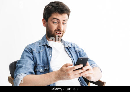 Ritratto di un bel scioccato uomo barbuto indossando abiti casual seduto nella sedia isolate su sfondo bianco, tenendo il telefono cellulare Foto Stock