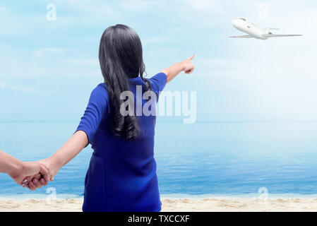 Vista posteriore della donna asiatica tenendo il suo fidanzato le mani sulla spiaggia e a puntare ad un piano di volo sopra il mare con cielo blu sullo sfondo Foto Stock