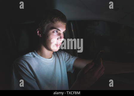 Distratti adolescente alla guida di una vettura con il suo telefono cellulare in mano. La luce dallo schermo del telefono è illuminare il suo volto. Foto Stock