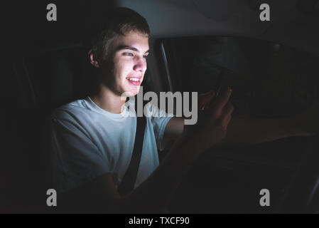 Distratti adolescente alla guida di una vettura con il suo telefono cellulare in mano. La luce dallo schermo del telefono è illuminare il suo volto. Foto Stock