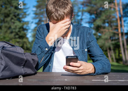 Interessato adolescente seduti ad un tavolo da picnic tutto solo con il suo telefono cellulare. Foto Stock