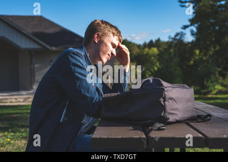 Sconvolto adolescente seduti ad un tavolo da picnic in un parco tutto da solo. Il suo zaino è sdraiato sul tavolo accanto a lui. Foto Stock