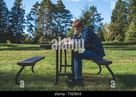 Interessato adolescente seduti ad un tavolo da picnic tutto solo con il suo telefono cellulare. Foto Stock