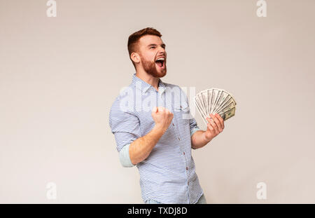 Inaspettata vittoria. Giovane ragazzo millenario che mostra sì segno con ventola di dollari in mano, panorama, spazio di copia Foto Stock