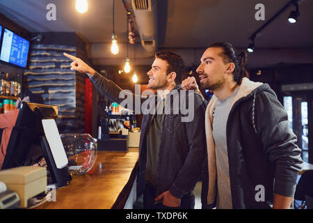 Vista laterale di due giovani uomini menu di lettura e la scelta di bevande mentre sta in piedi vicino al contatore nel pub moderno Foto Stock