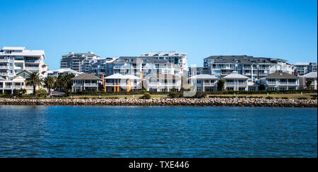 Harbourside appartamenti di un condominio dietro free standing case con facciata di erba, pietra parete di ritegno e il fiume azzurro contro il cielo blu Foto Stock