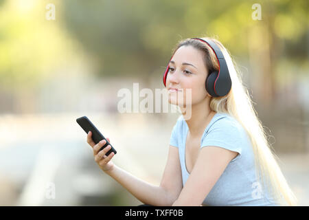 Ragazza rilassati ascoltando la musica con le cuffie e smart phone che guarda lontano seduto in un parco Foto Stock