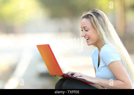 Vista laterale verticale di un felice ragazza adolescente usando un computer portatile rosso sititng in un parco Foto Stock