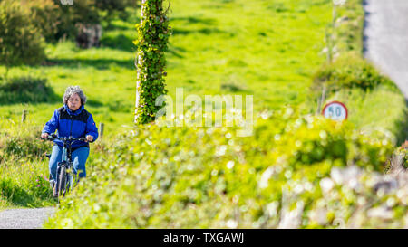 Stanco matura donna messicana cavalcare la sua bicicletta tra i prati di una collina, espressione sul viso, aria espirata, splendida soleggiata giornata di primavera in vill Foto Stock