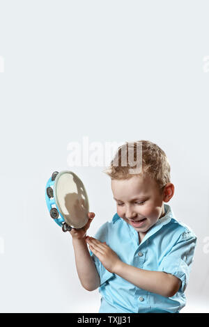 Allegro ragazzo in una maglietta blu tenendo un tamburello e sorridente su uno sfondo luminoso Foto Stock