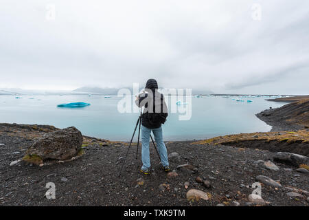 Giovane fotografo in inverno freddo rivestire tenendo immagine fotografia fotografare ghiacciaio blu iceberg nel lago laguna in Islanda su treppiede Foto Stock