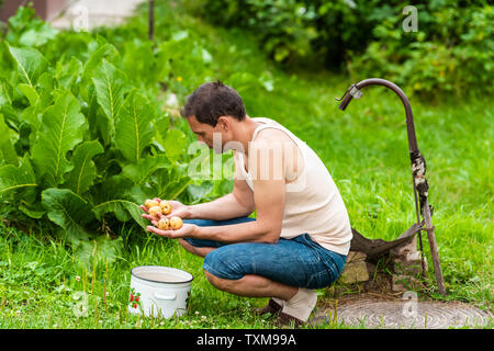 Giovane agricoltore il risciacquo lo sporco da homegrown patate in estate verde in Ucraina seduto da foglie di rafano pianta, ben rubinetto il lavaggio della verdura Foto Stock