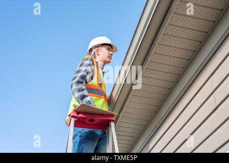 Home inspector in piedi su una scaletta e fornendo una ispezione al tetto di una casa. Foto Stock