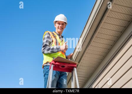 Home inspector in piedi su una scaletta e fornendo una ispezione al tetto di una casa. Foto Stock