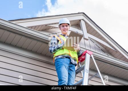 Giovane architetto maschio in piedi su una scaletta e vestito di ingranaggio di sicurezza. Foto Stock